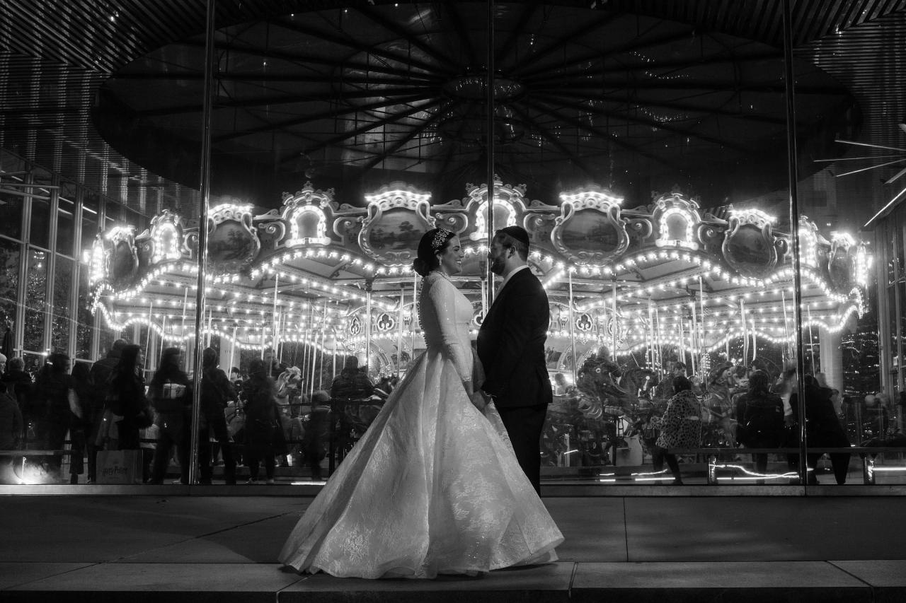 Black-and-white wedding photo of a bride and groom holding hands walking forward together at an illuminated carousel-style event with a crowd behind them.
