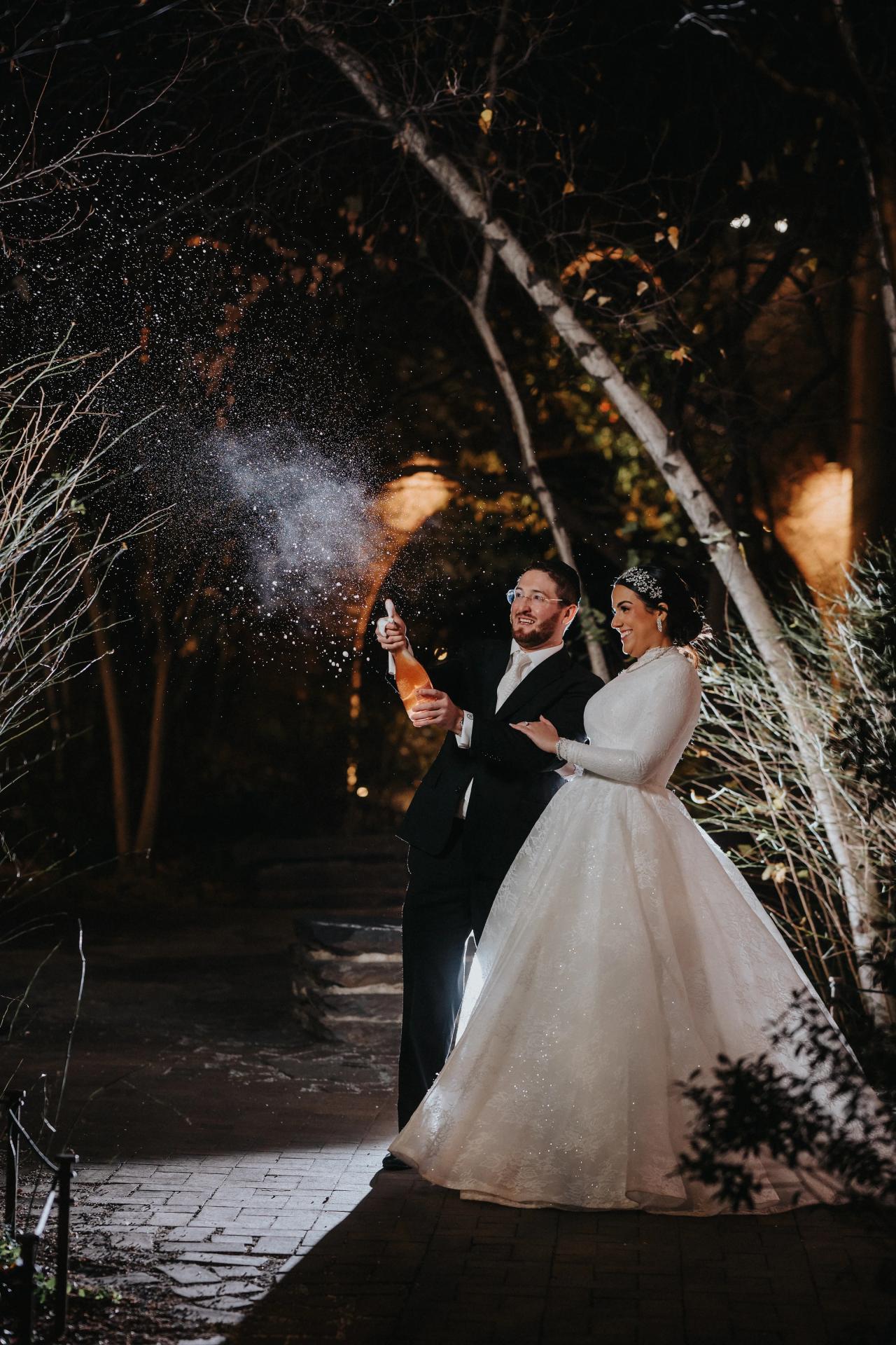 Frum bride and groom smiling and walking together with confetti/spark particles bursting in the air at night, warm backlight, branches framing the scene.