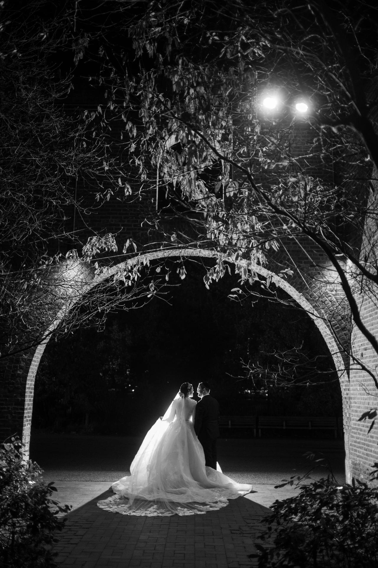 Black-and-white nighttime wedding portrait of a bride and groom framed by an arched brick passageway and branches, with a bright light halo behind them and the bride’s veil flowing.