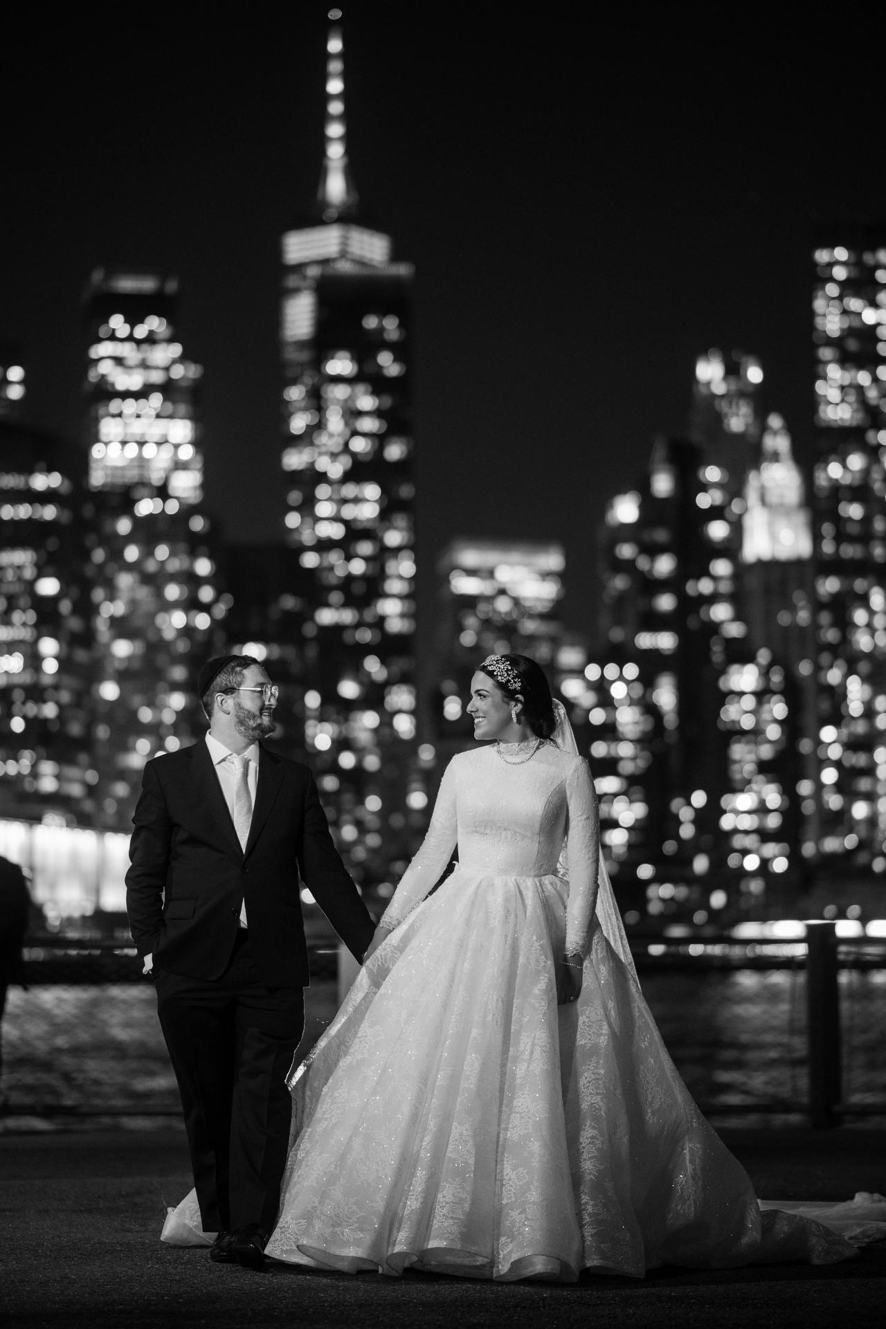 Black-and-white skyline wedding portrait of a bride and groom holding hands and walking at night, with glowing city lights bokeh in the background.