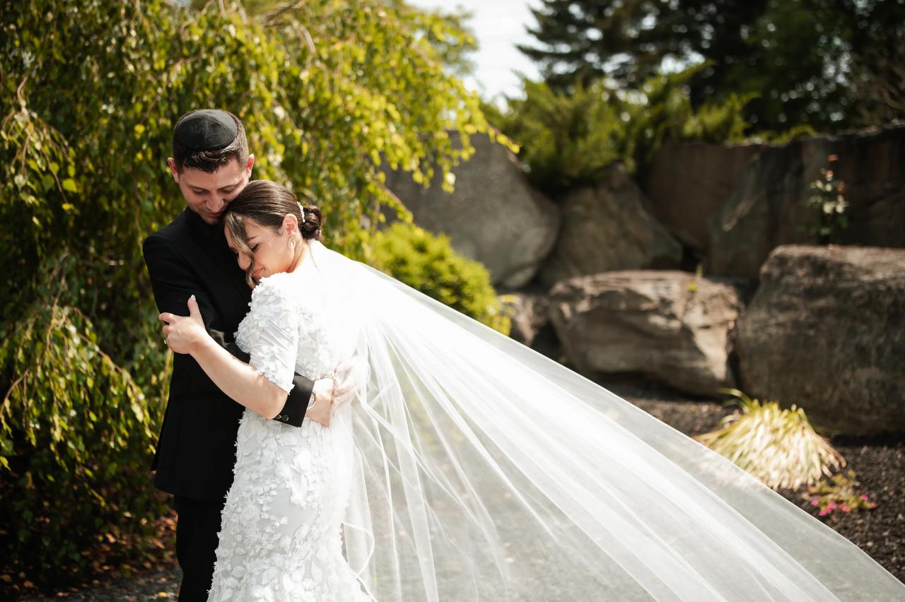 Groom holding the bride in an intimate embrace outdoors in warm sunlight; her white veil flows across the frame with greenery and a stone wall behind them. Wedding photography at The Barn At Indian Ridge, Highland, NY.
