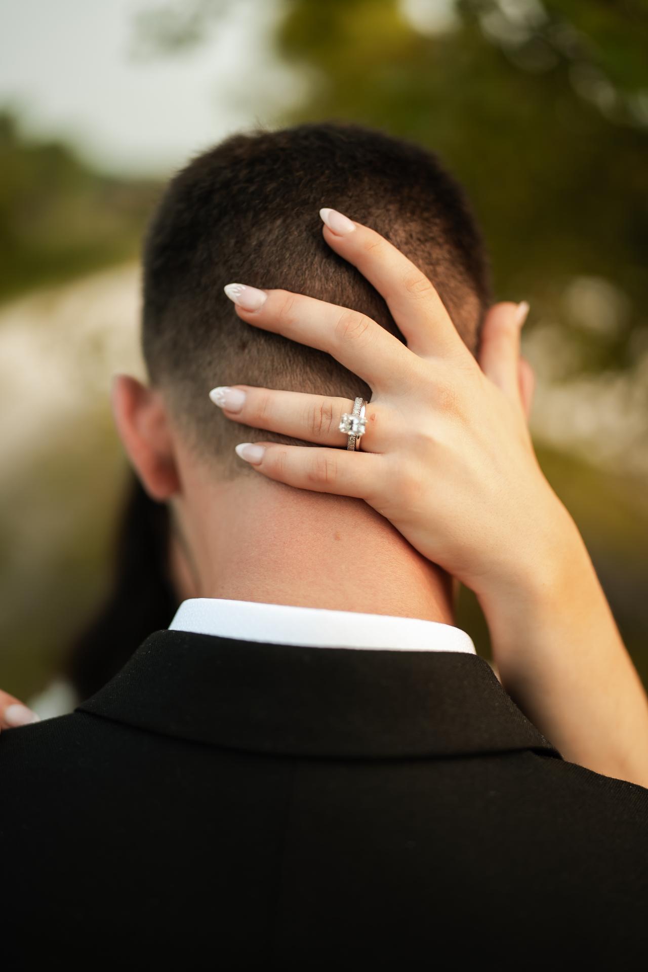 Close detail of the bride’s ringed hand cupping the back of the groom’s head during an embrace, with soft green garden bokeh. Wedding photography at The Barn At Indian Ridge, Highland, NY.