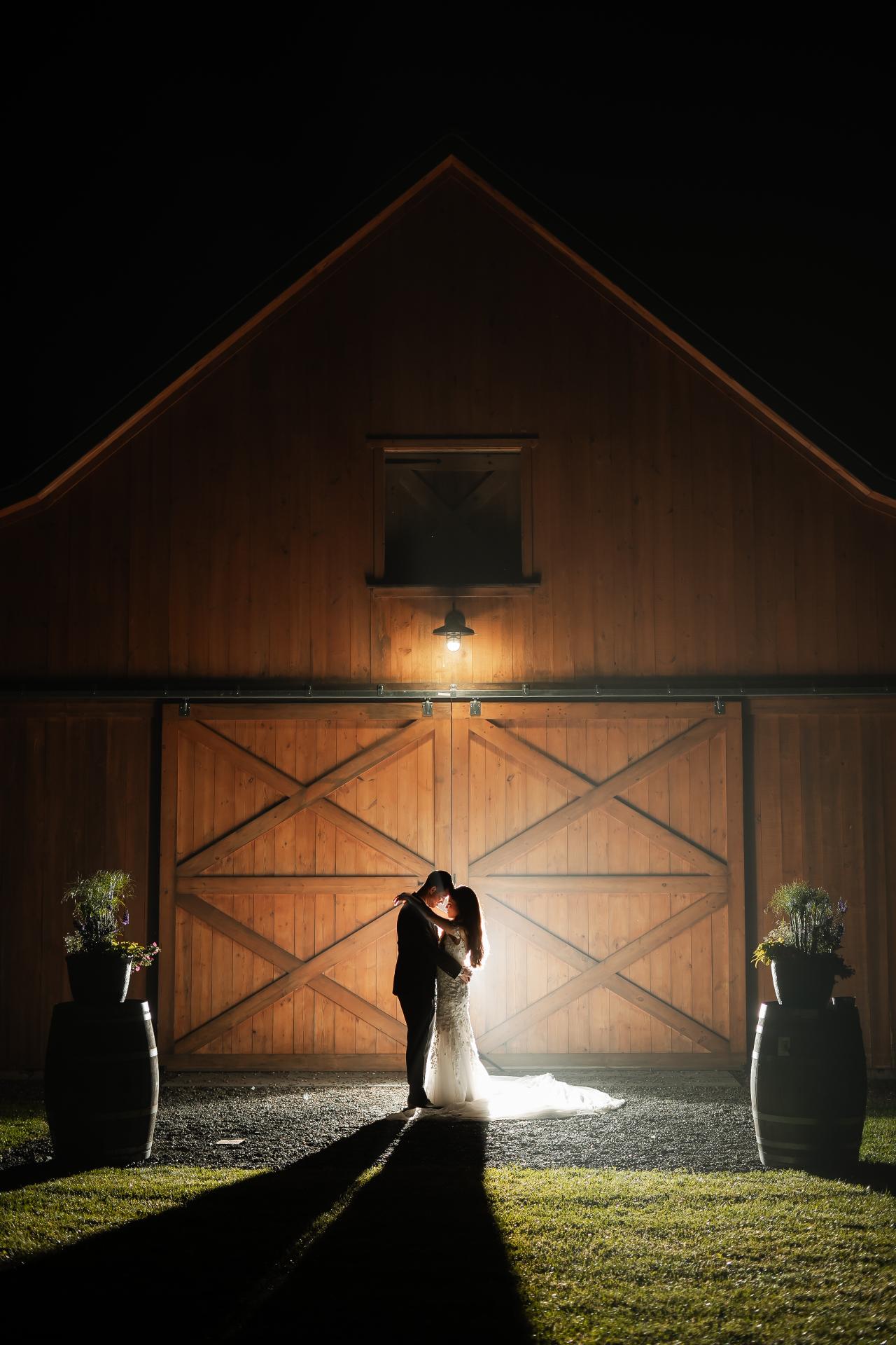 Framed by a wooden barn entrance, the bride and groom share a kiss in strong directional light with long shadows on the grass. Wedding photography at The Barn At Indian Ridge, Highland, NY.