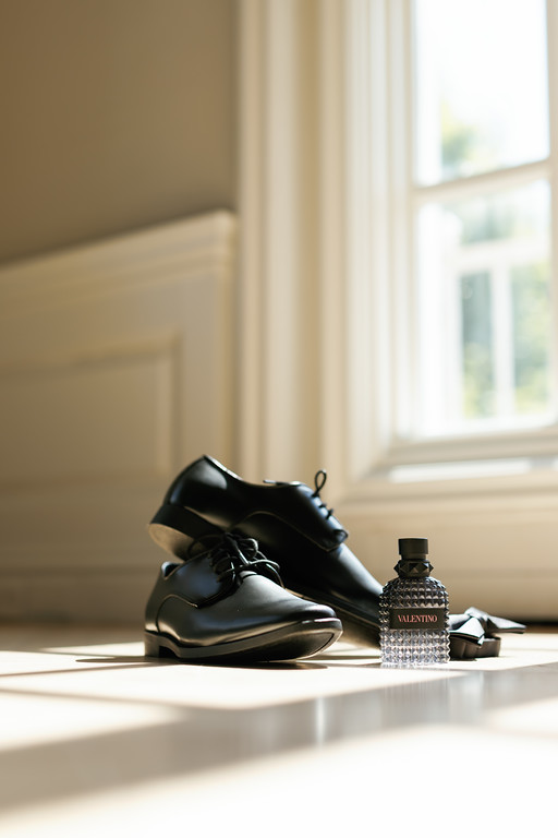 Black dress shoe and a Valentino cologne bottle arranged together on a bright windowsill with soft natural light.