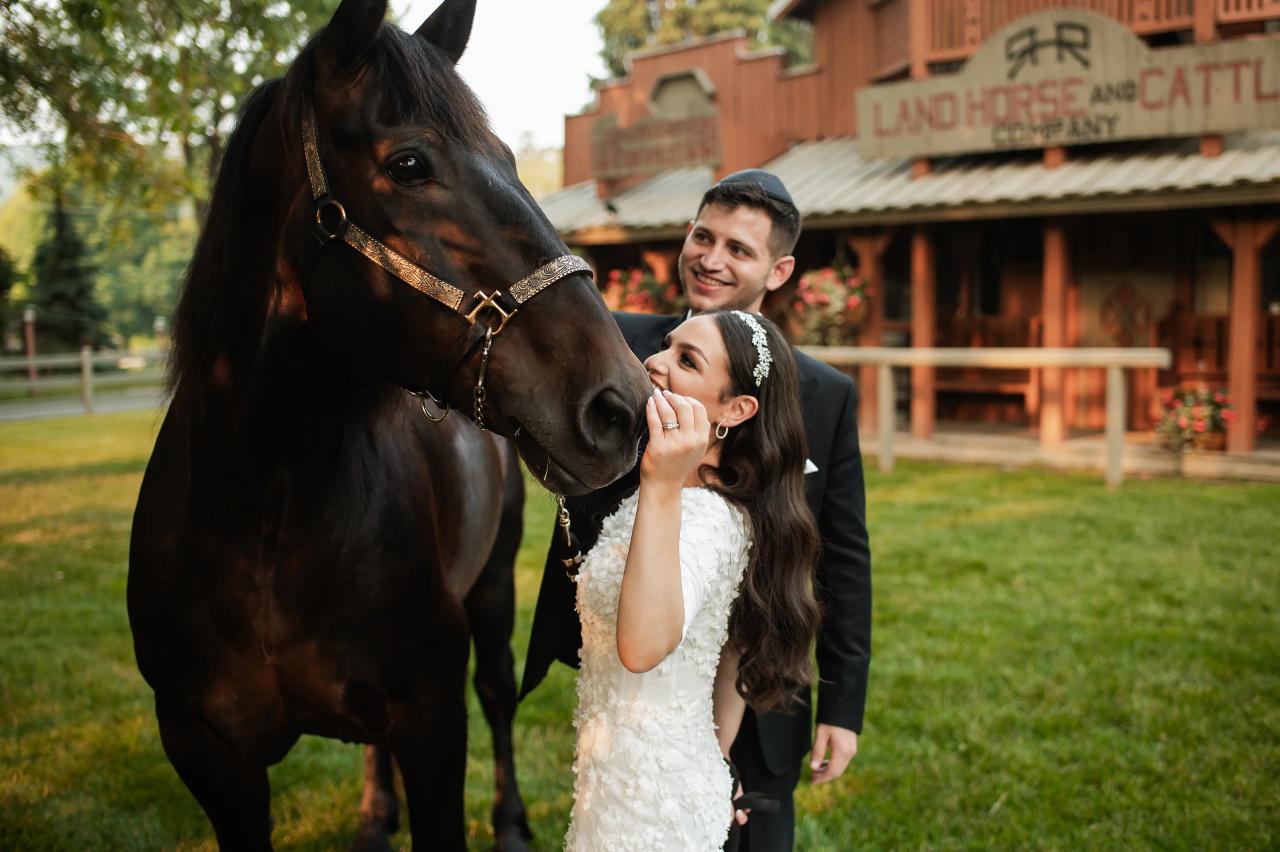 The bride leans toward a black horse in a playful garden portrait, with the smiling groom beside her and a rustic building behind them. Wedding photography at The Barn At Indian Ridge, Highland, NY.
