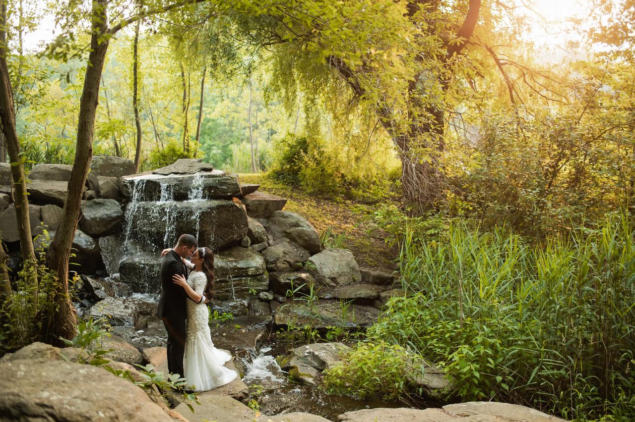 Couple embracing and kissing beside a stone waterfall in a lush outdoor setting at golden hour, with warm sunlight filtering through trees. Wedding photography at The Barn At Indian Ridge, Highland, NY.