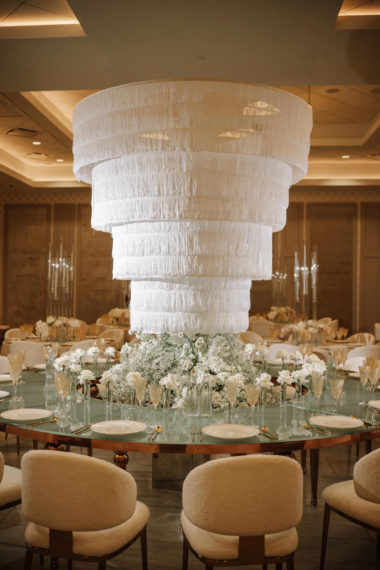Warmly lit reception hall with a tall white textured centerpiece tower above a floral table display, chairs and glassware surrounding it. Bar Mitzvah photography at Hilton Garden Inn Springfield, Springfield, NJ.