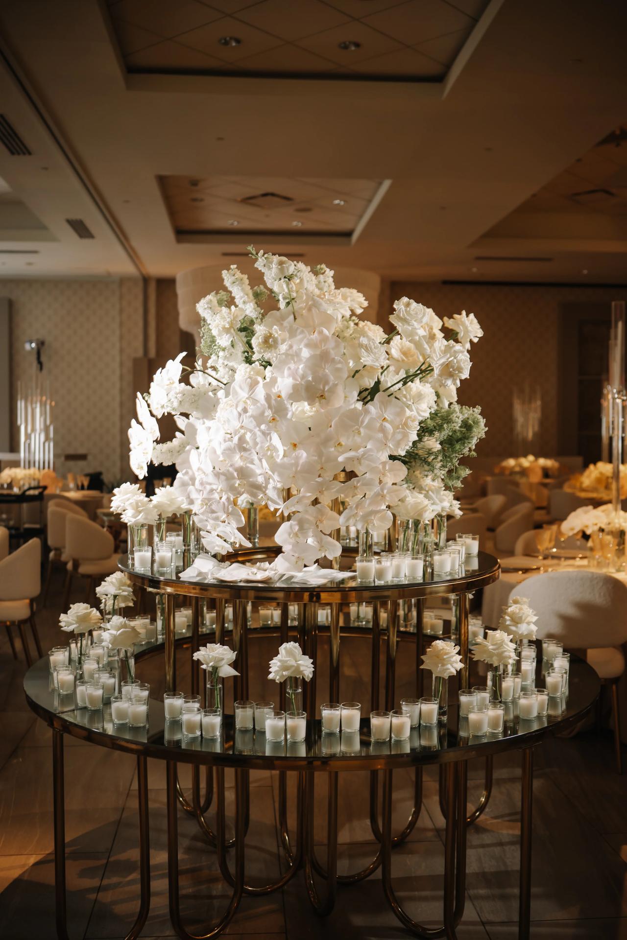 Round event display table with abundant white floral arrangement and many small votive candles in a warm-lit ballroom. Bar Mitzvah photography at Hilton Garden Inn Springfield, Springfield, NJ.
