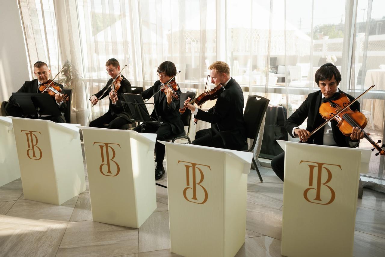 Violinists seated and performing at music stands behind illuminated panels near large windows. Bar Mitzvah photography at Hilton Garden Inn Springfield, Springfield, NJ.