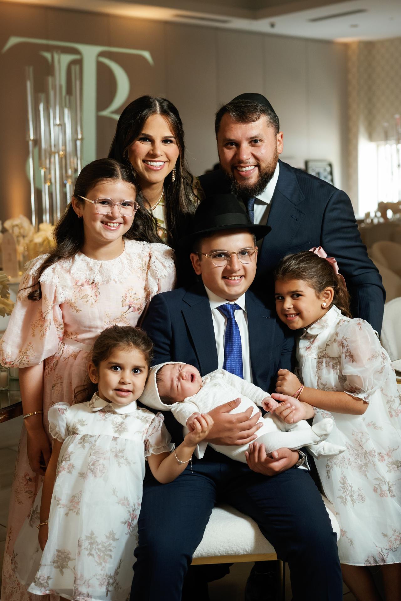 Smiling couple holding a newborn with several children in coordinated white outfits during a formal family portrait indoors. Bar Mitzvah photography at Hilton Garden Inn Springfield, Springfield, NJ.