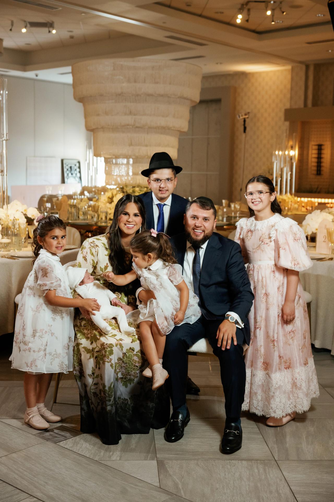 A family group with adults and children in coordinated formalwear poses against a warmly lit ballroom backdrop. Bar Mitzvah photography at Hilton Garden Inn Springfield, Springfield, NJ.