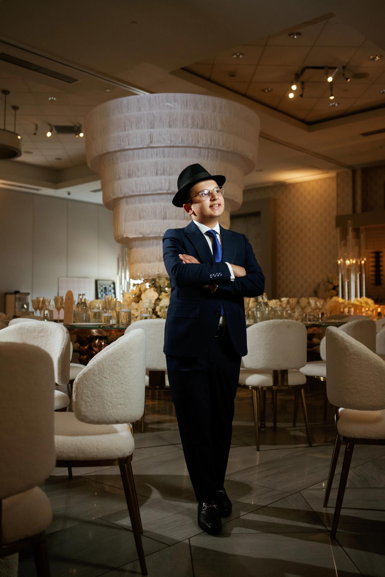Smiling young boy in a navy suit and hat with arms crossed, standing in a warmly lit banquet hall with a large centerpiece behind him. Bar Mitzvah photography at Hilton Garden Inn Springfield, Springfield, NJ.