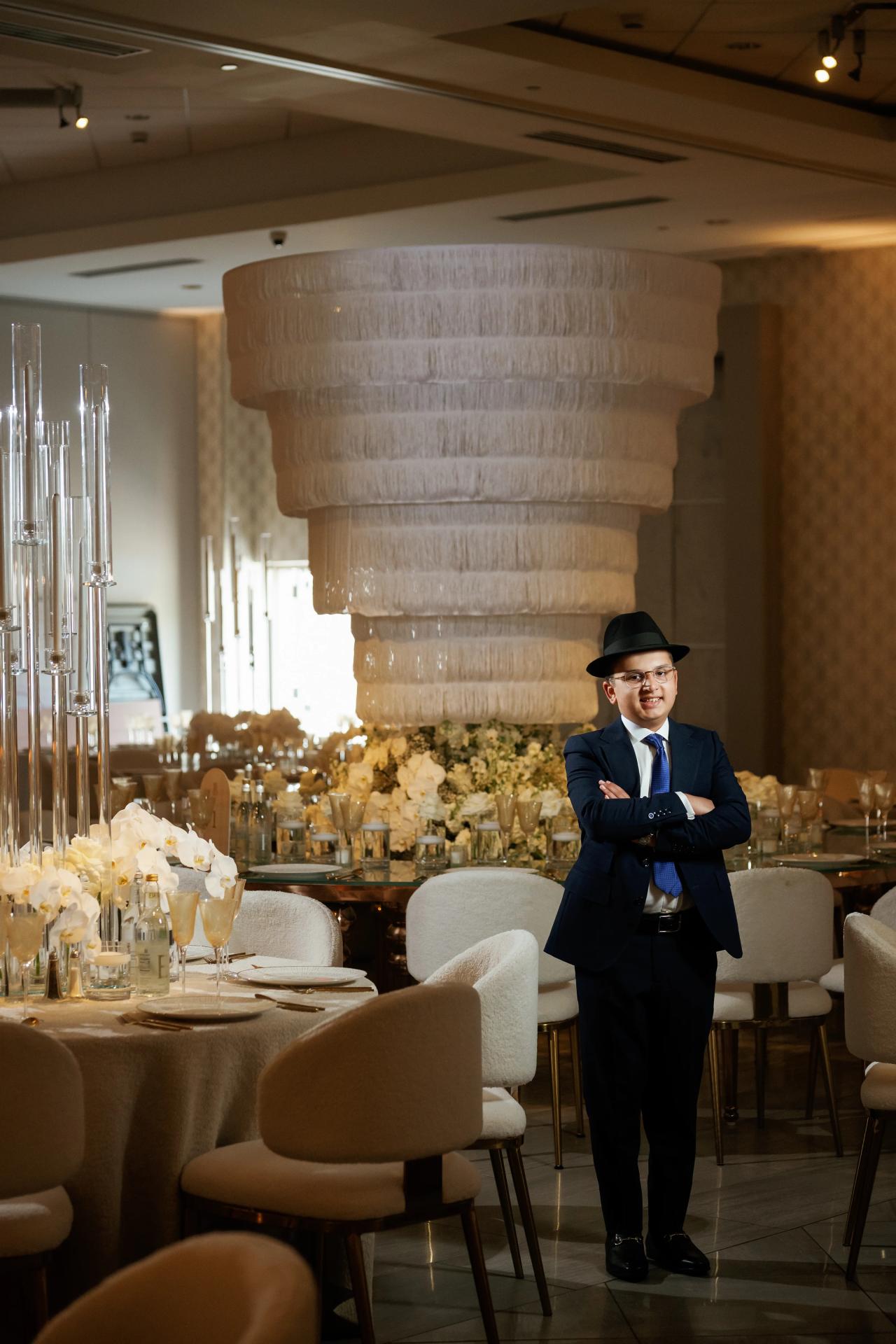A boy in a black hat and formal suit stands with arms crossed in a banquet hall in front of a tall tiered floral centerpiece. Bar Mitzvah photography at Hilton Garden Inn Springfield, Springfield, NJ.
