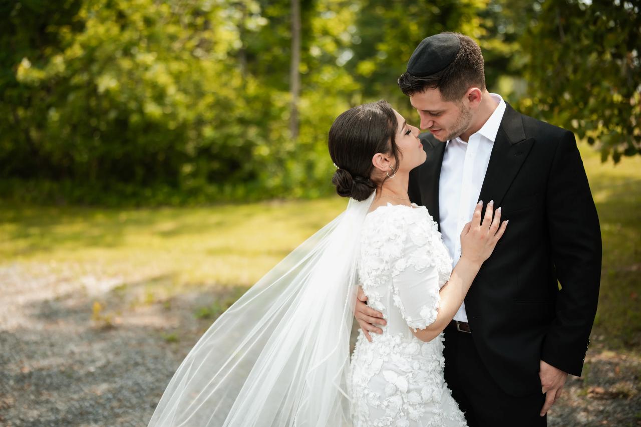Bride and groom in close embrace outdoors, gazing at each other with soft smiles and hands held near the couple’s faces during the badeken, with lush greenery in the background. Wedding photography at The Barn At Indian Ridge, Highland, NY.