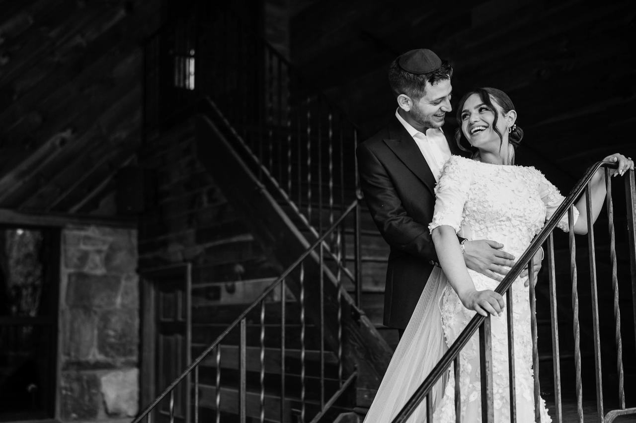 Black-and-white photo of a bride and groom laughing together on a staircase, framed by a dark architectural backdrop and iron railing. Wedding photography at The Barn At Indian Ridge, Highland, NY.