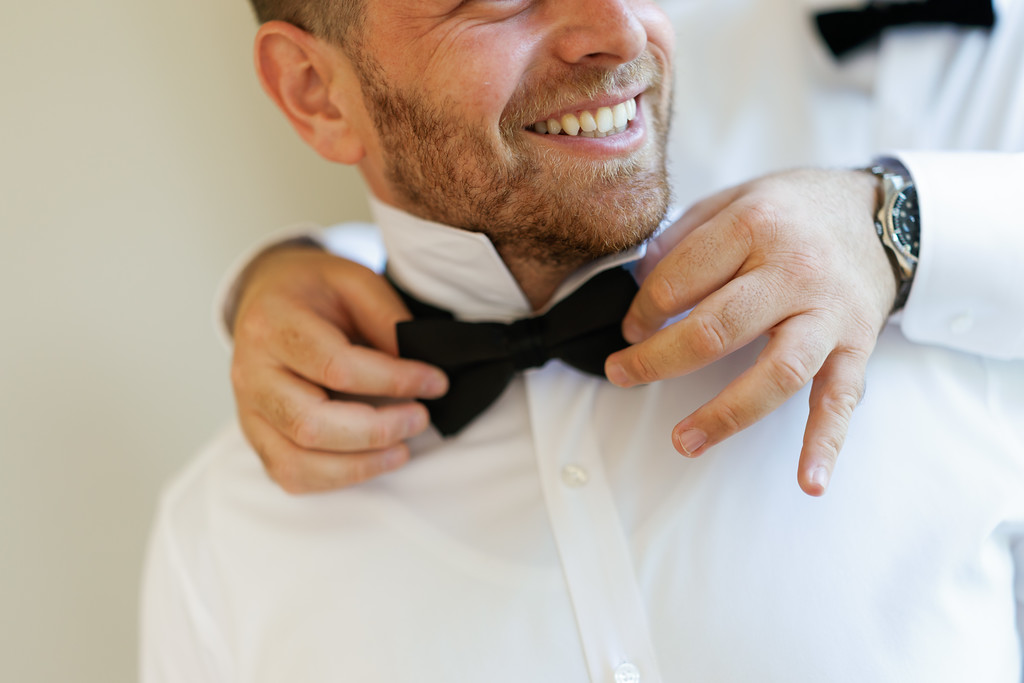 Smiling groom adjusting his bow tie while buttoning his white shirt, hands near his collar during wedding getting-ready.