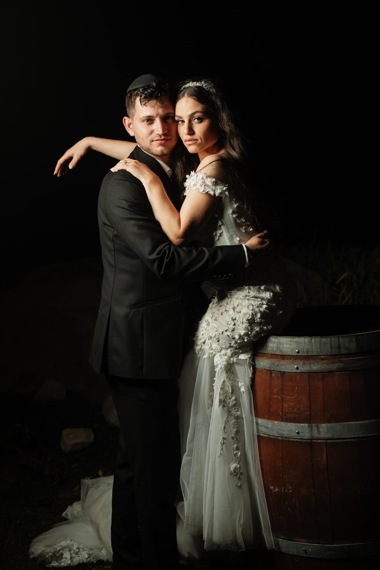 Bride and groom in a close embrace during the celebration, lit dramatically against a dark background with the bride’s ornate dress visible and a wooden barrel in the foreground. Wedding photography at The Barn At Indian Ridge, Highland, NY.