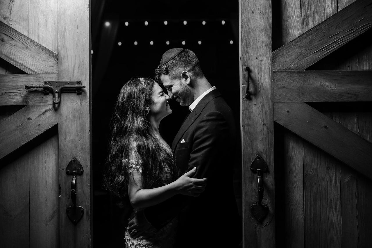 Black-and-white close portrait of a bride and groom sharing an intimate forehead kiss while framed by dark wooden door panels. Wedding photography at The Barn At Indian Ridge, Highland, NY.