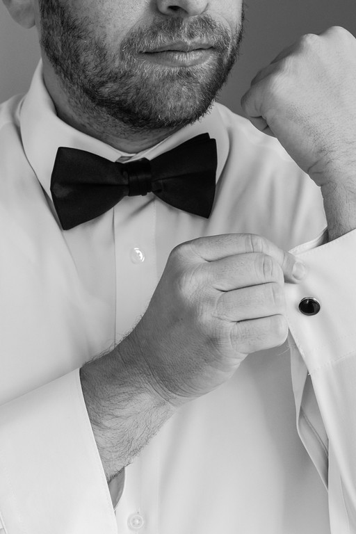 Black-and-white close-up portrait of a man adjusting his black bow tie and cufflinks, hands near his shirt collar.