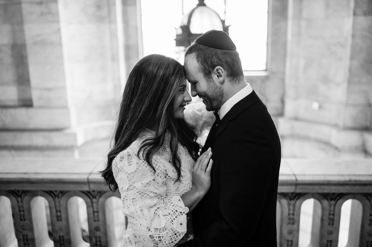 Black-and-white portrait of a bride and groom in close embrace, leaning their foreheads together in a grand stone hall with soft window light.
