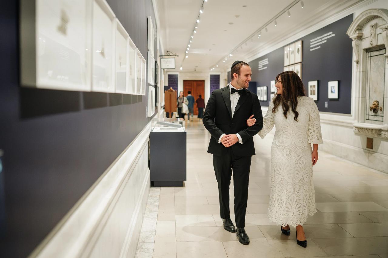 Bride and groom walking together through an exhibition corridor at the New York Public Library, lined with artwork displays.