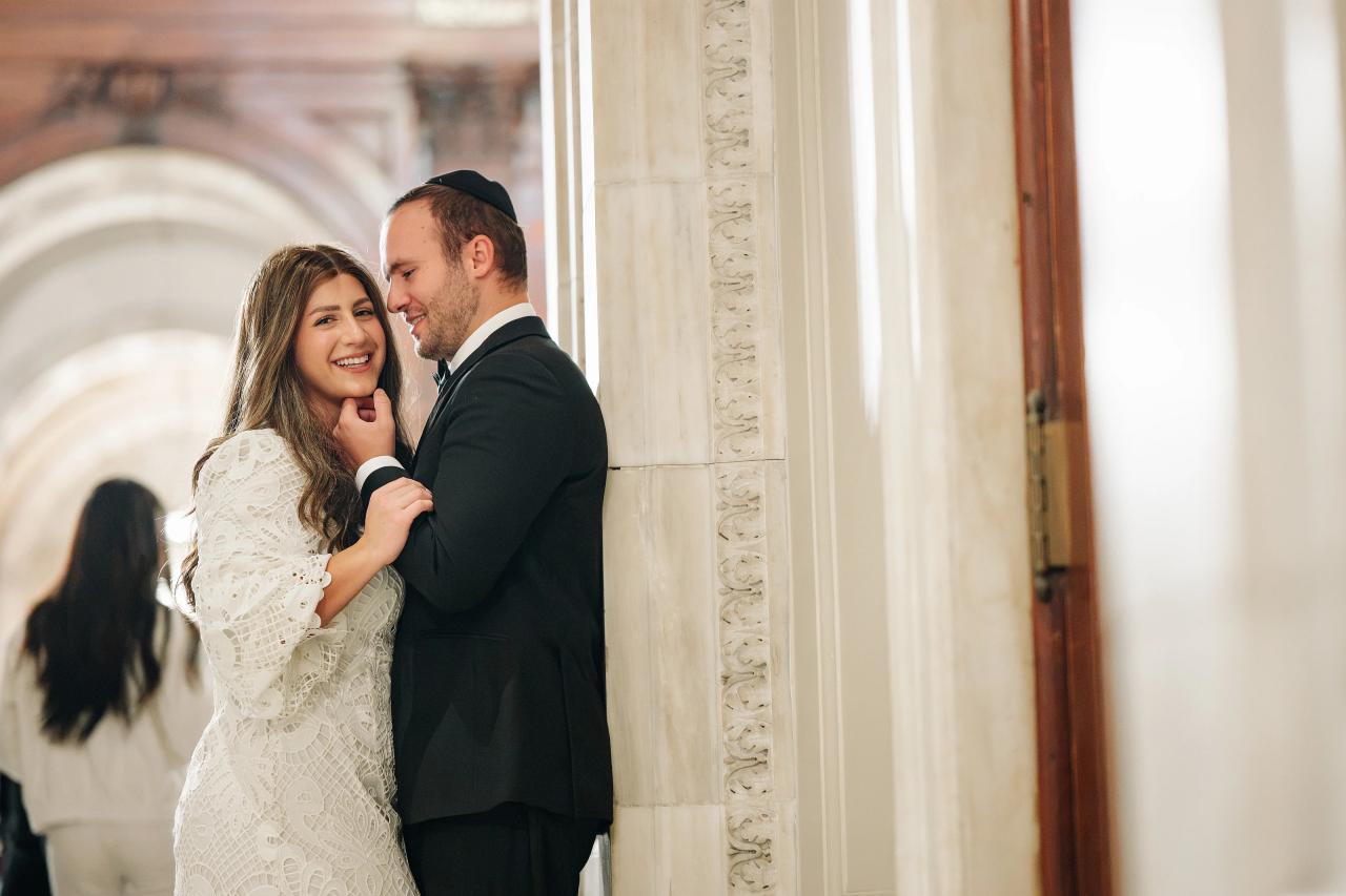 Bride and groom smiling close together indoors by tall stone columns and an arched doorway, warm soft light, intimate candid portrait.