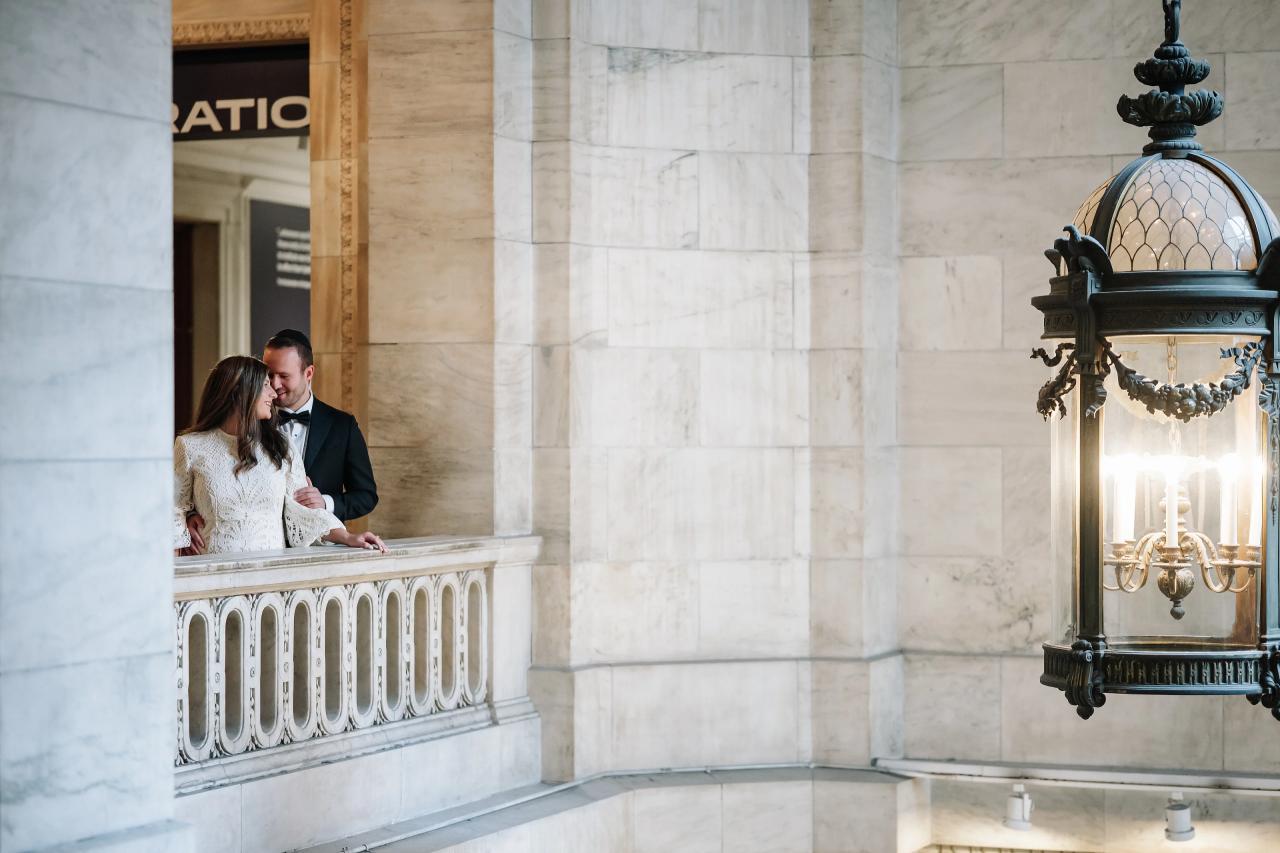 Couple sharing an intimate moment at a balcony railing inside the New York Public Library, with warm window light.