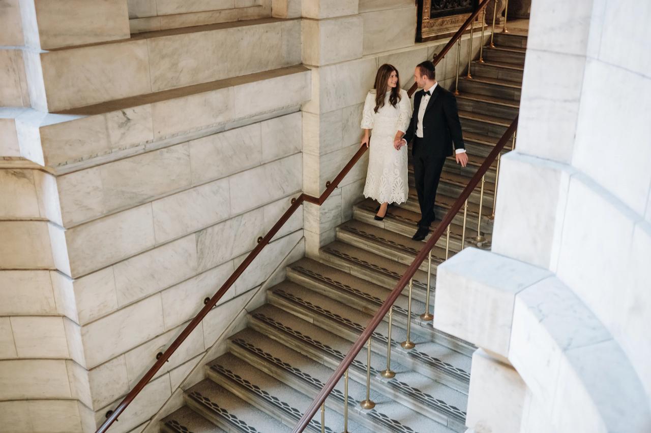 Wedding couple in coordinated attire walking together on a grand marble stair landing inside a stone building, framed by banisters and columns.