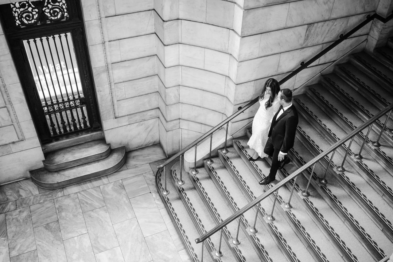 Black-and-white wedding portrait of a bride and groom walking down a grand staircase inside a large stone building, framed by railings and architectural columns.