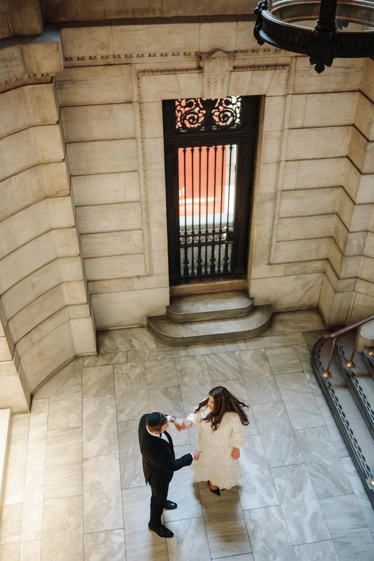 Overhead view of a couple in formal attire on a stone-tiled floor inside a grand building, with a decorative arched doorway in the background.