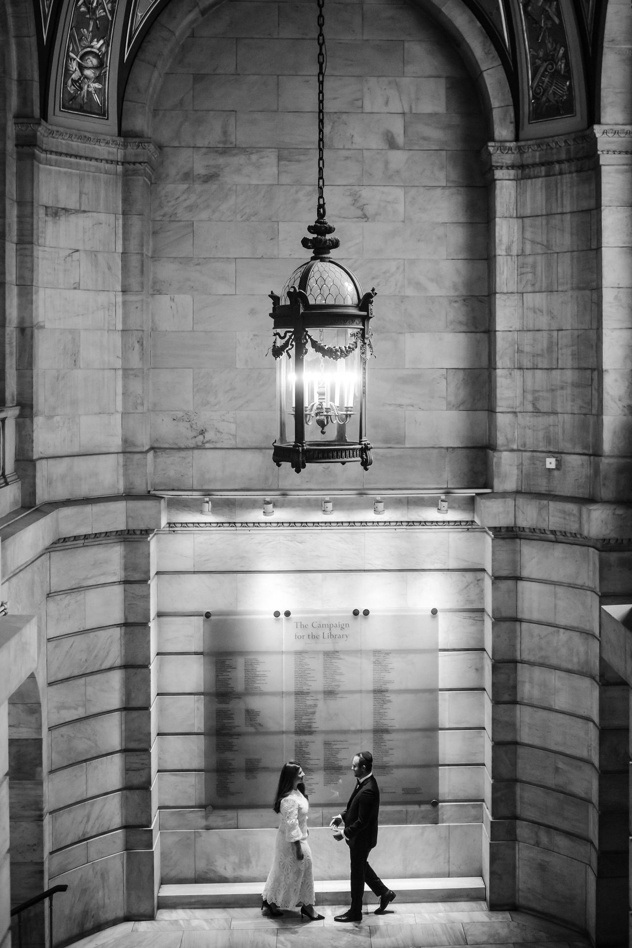 Black-and-white photo of a bride and groom walking together under a glowing hanging lantern inside a stone-arched building.
