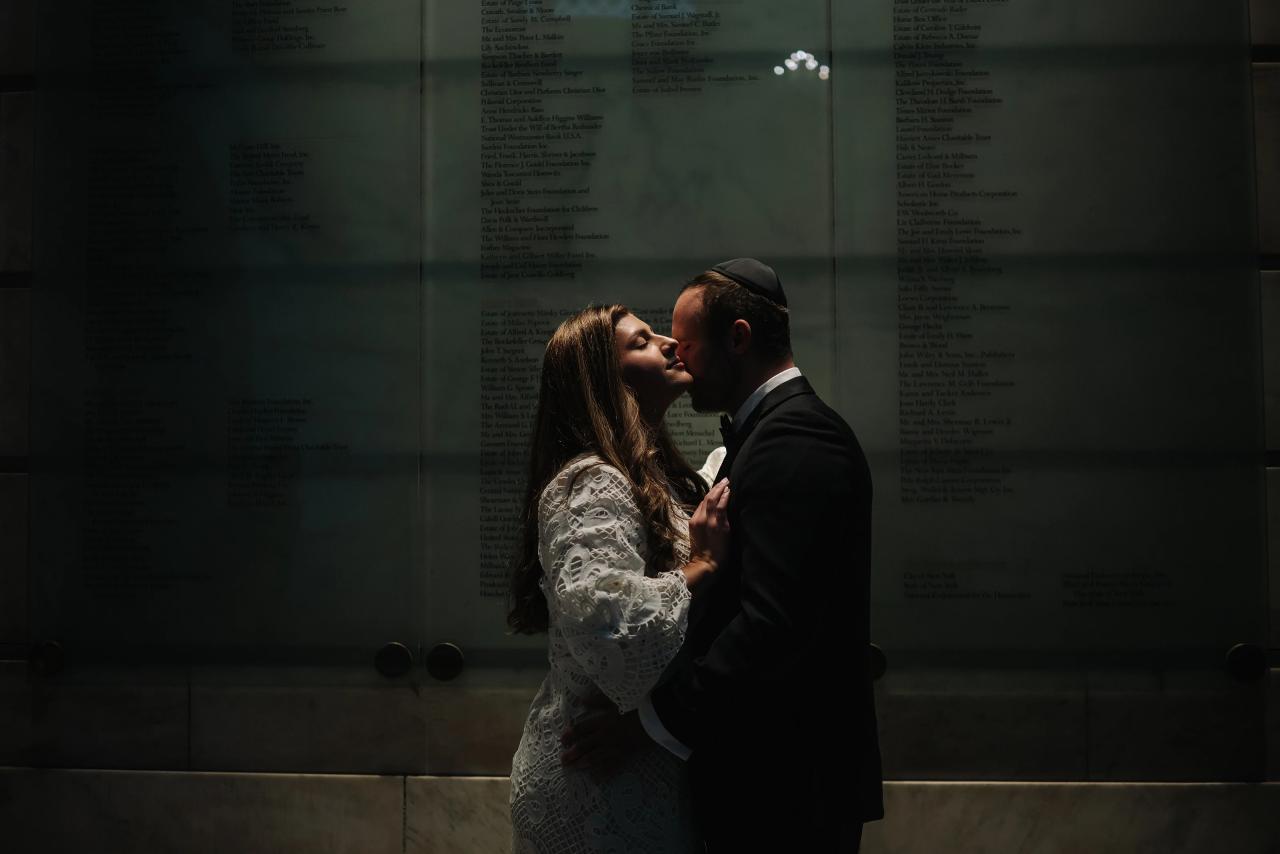Couple kissing in a dark, moody interior with stone walls and text panels behind them; the bride wears a white lace dress and the groom wears a black suit and kippah.