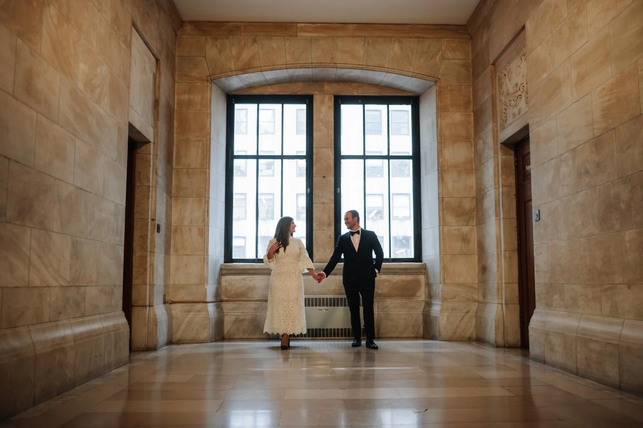 Bride and groom holding hands in a grand stone interior, framed by bright backlit windows through a doorway.