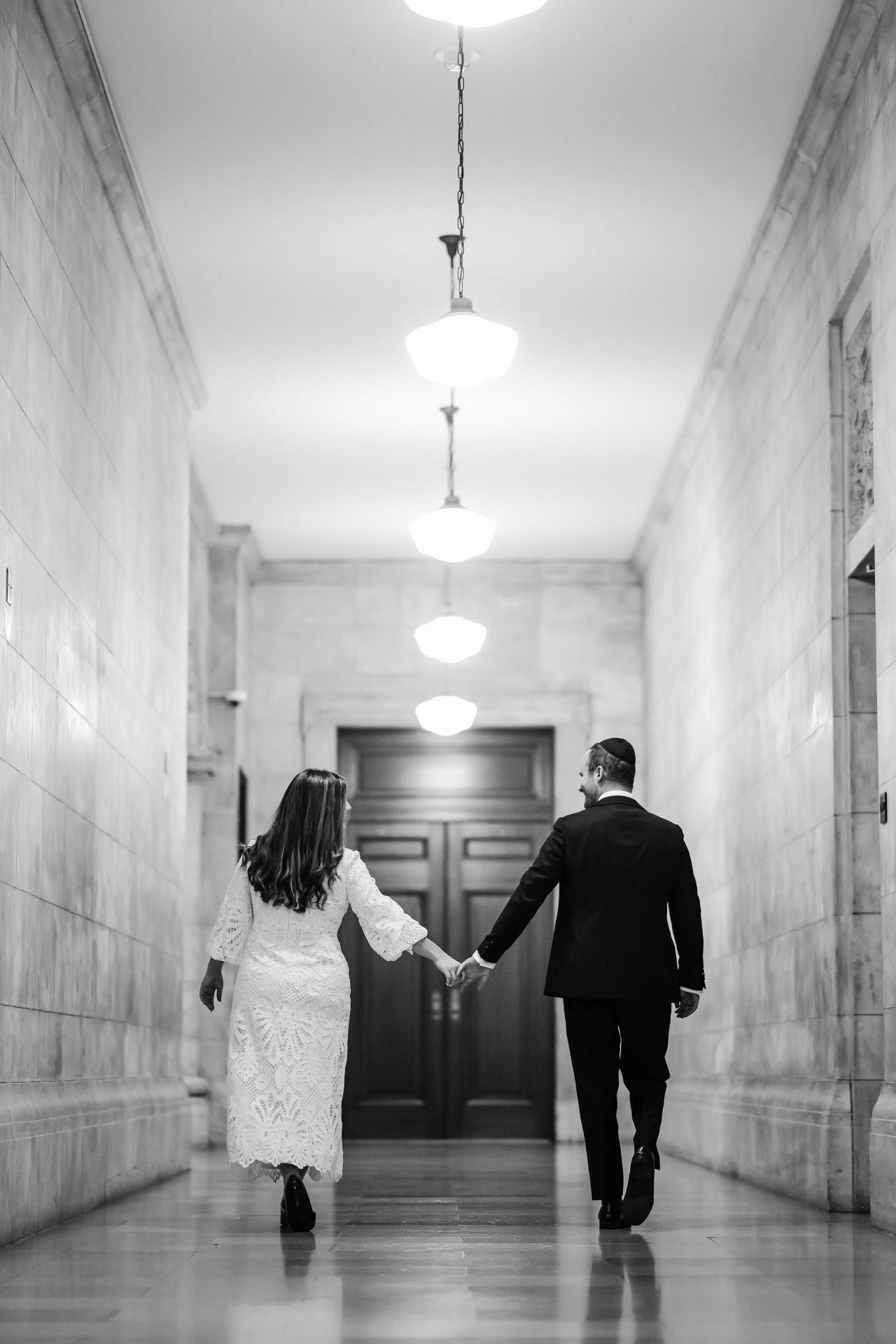 Black-and-white wedding portrait of a couple holding hands walking down a long corridor with repeating overhead lights and reflective floor.