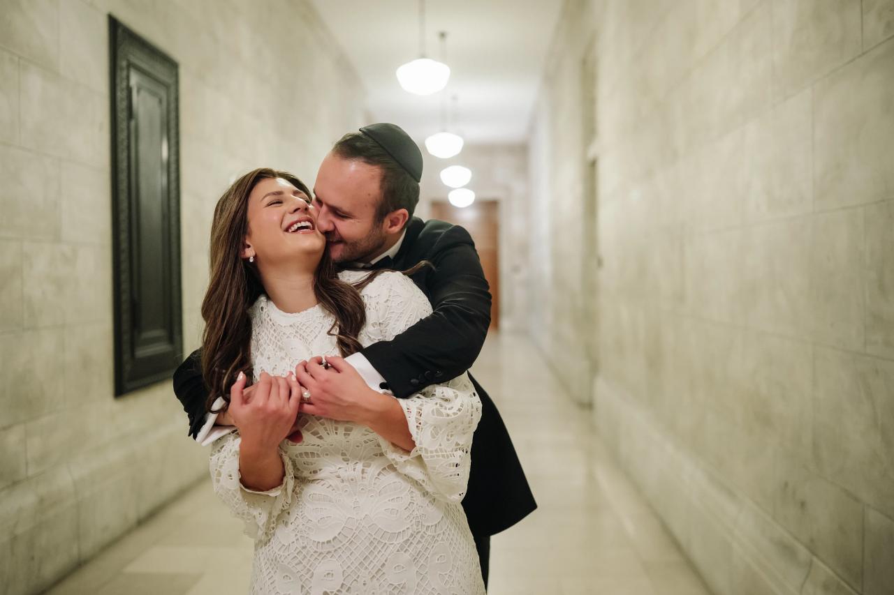 Wedding couple laughing and embracing in a bright marble corridor, bride in a white lace dress and groom in a dark suit and cap.