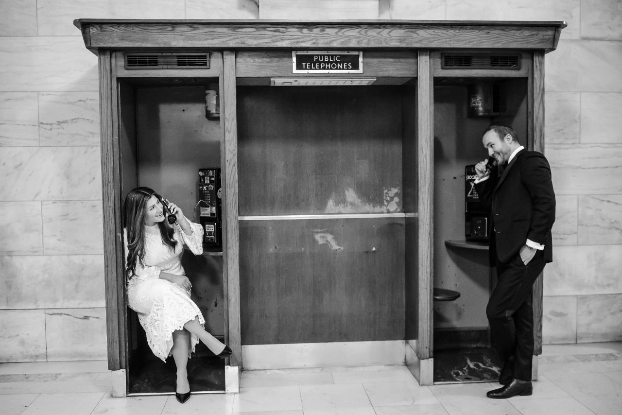 Black-and-white wedding candid: bride leaning out from a phone booth while the groom stands beside her, both smiling, with a “PUBLIC TELEPHONES” sign overhead.