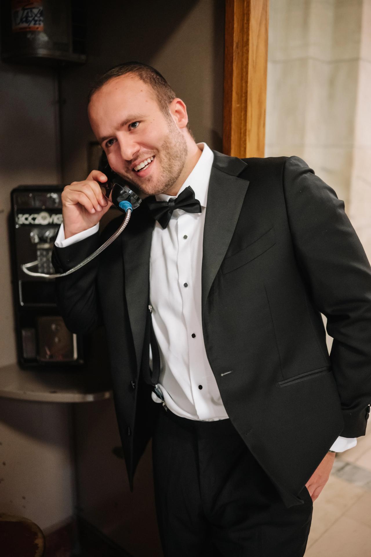 Groom in a black tuxedo smiling while holding a vintage telephone receiver at the New York Public Library.