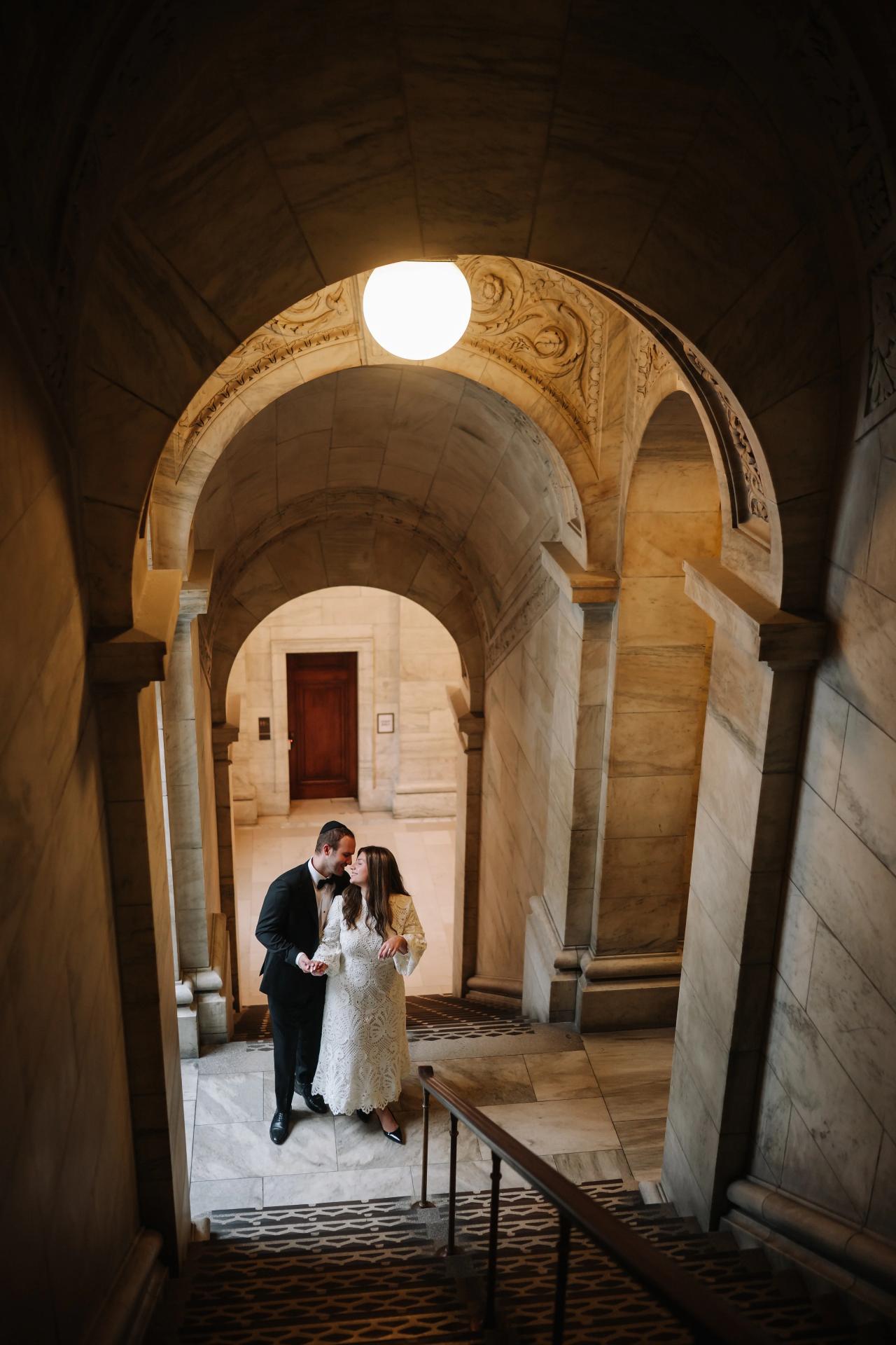 Bride and groom walking closely together inside a grand stone archway corridor under a warm overhead light.