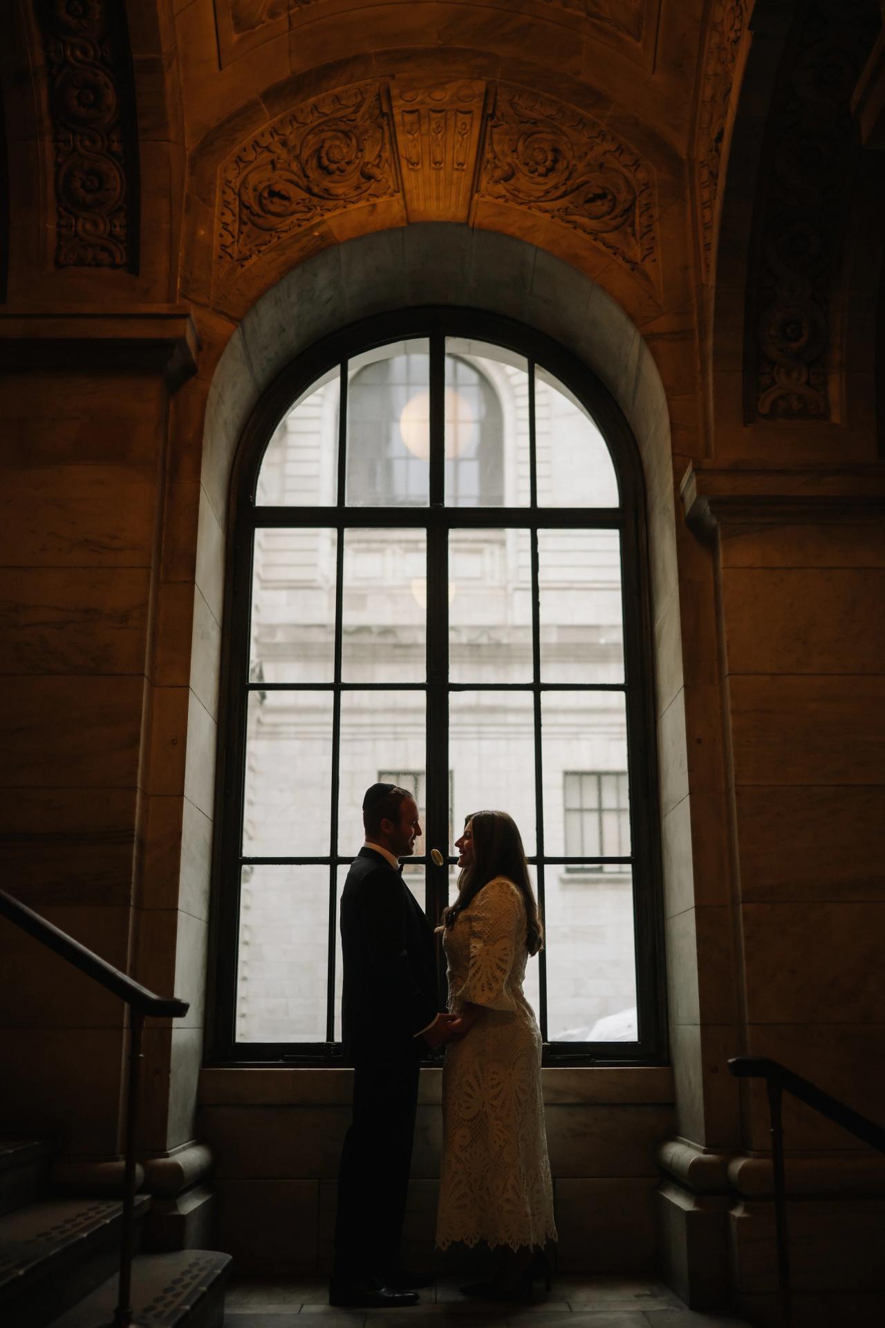 Silhouetted couple facing one another at the New York Public Library, framed by a tall arched window and warm stonework.