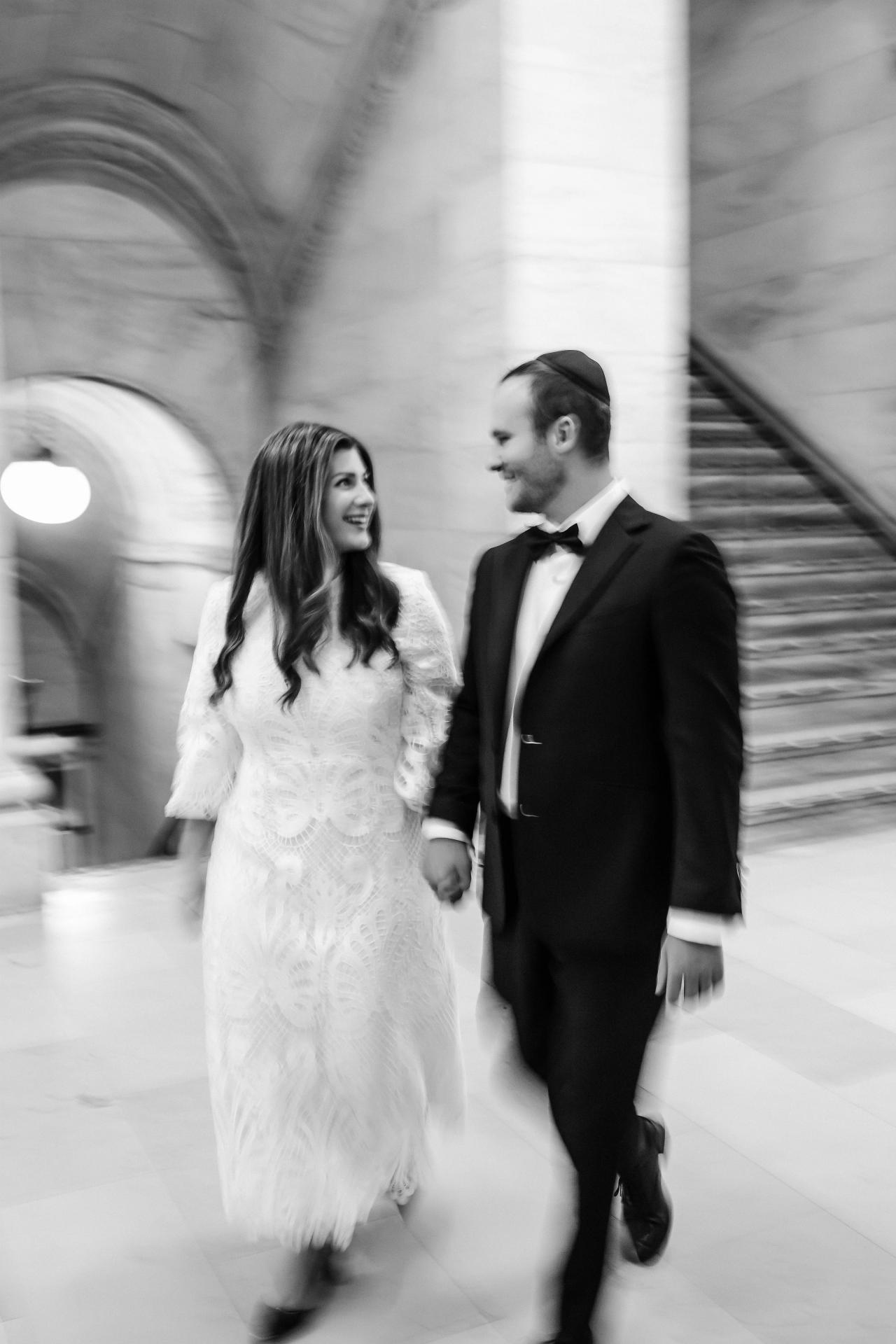 Black-and-white photo of a bride and groom walking together indoors in a hallway, with motion blur in the background and a softly smiling, side-profile moment.