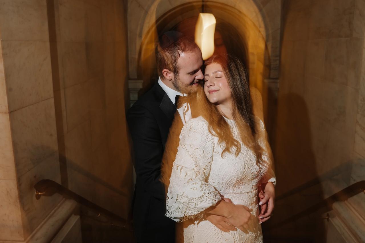 Wedding couple in warm golden light kissing in an elegant stone corridor, bride in lace dress and groom in tuxedo embracing closely.