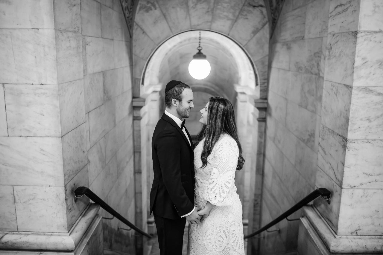 Black-and-white wedding photo of a bride and groom holding hands inside a stone archway corridor, looking at each other under a bright central light.
