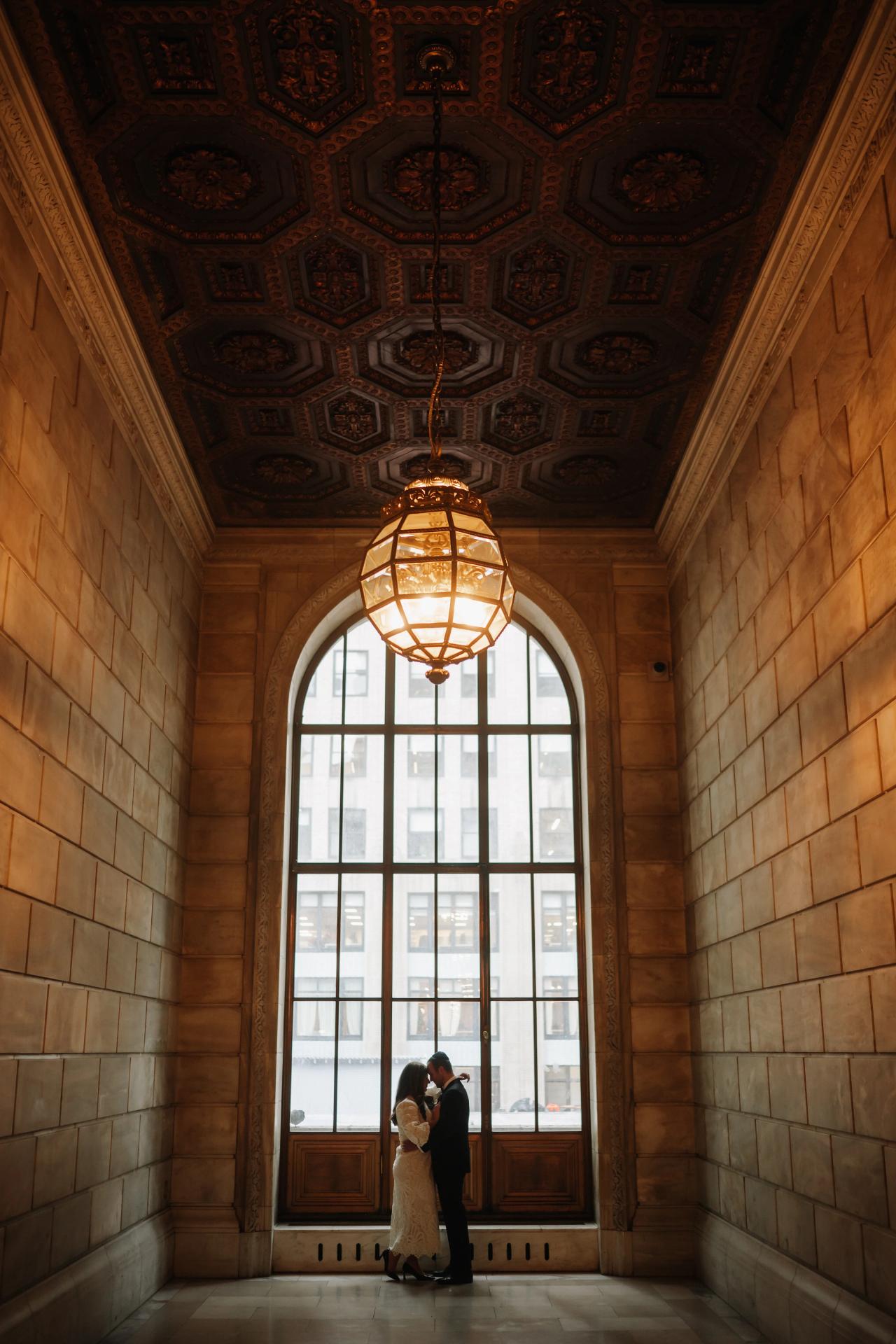 Bride and groom share an intimate kiss in a warmly lit arched corridor with a glowing chandelier and tall window behind them.