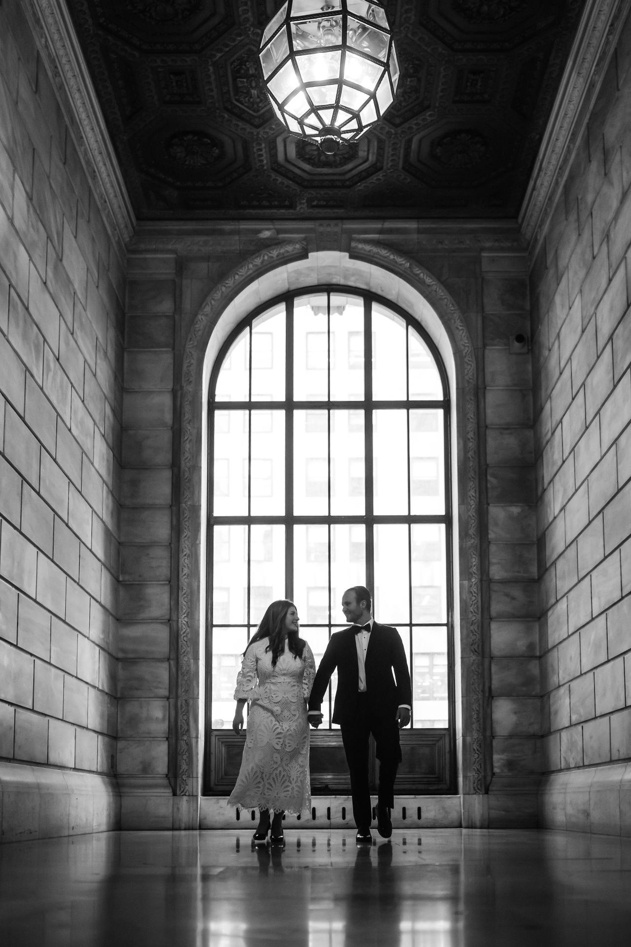 Black and white wedding portrait of a couple walking hand-in-hand toward a bright arched window inside a grand hall.