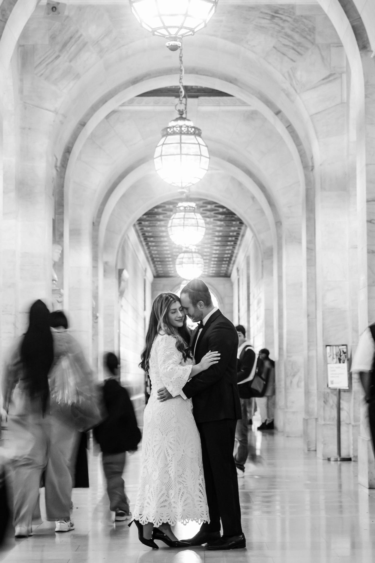 Black-and-white wedding photo of a bride and groom sharing a kiss while walking in a long arched corridor, with chandelier lights and softly blurred guests behind them.