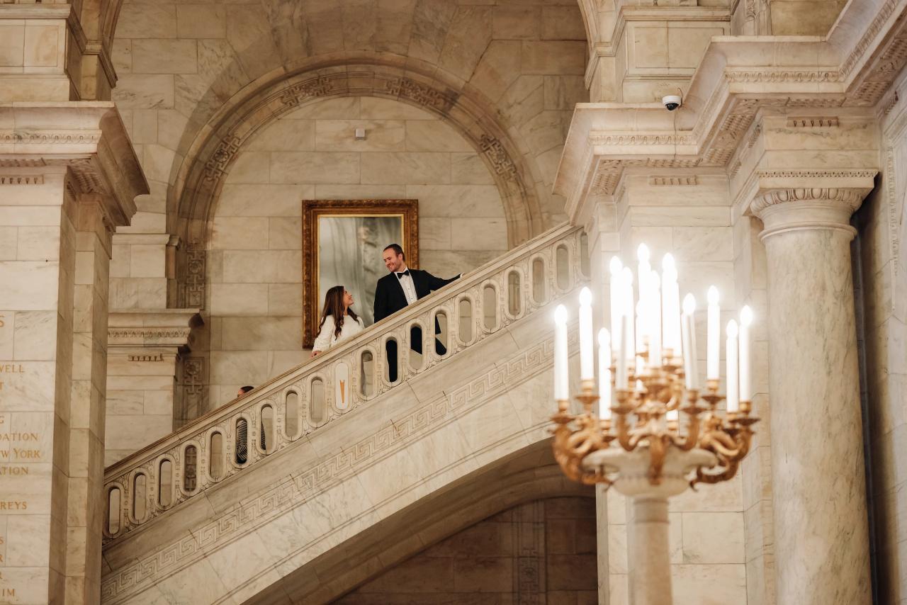 Bride and groom kissing in a warm-lit grand stair hall with marble arches and a chandelier in the foreground.
