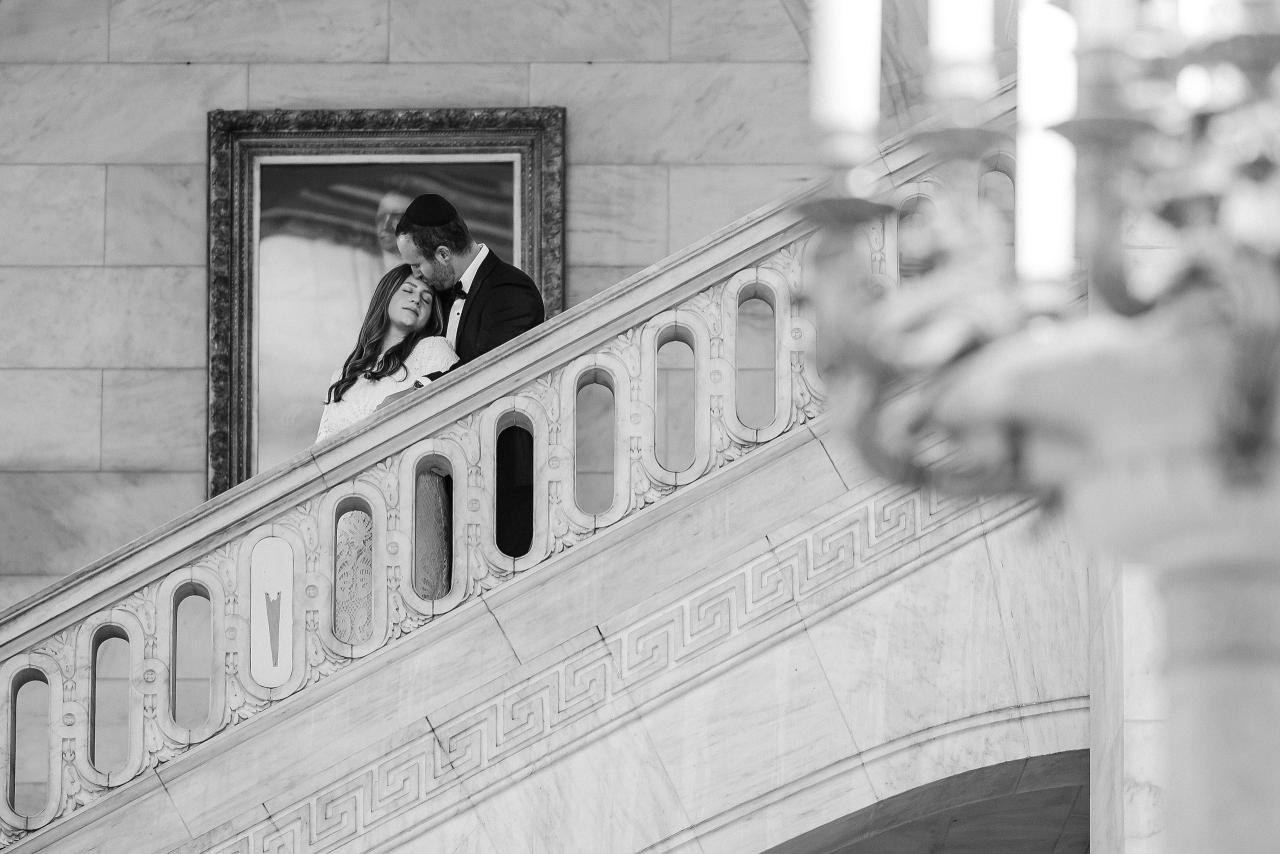 Black-and-white wedding portrait of a couple on an ornate balcony, leaning in close and sharing a quiet embrace, with architectural details in the foreground.