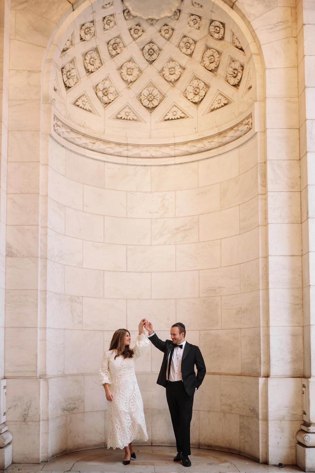 Bride and groom in matching formalwear standing by a cream stone archway, facing each other and looking up with a celebratory gesture.