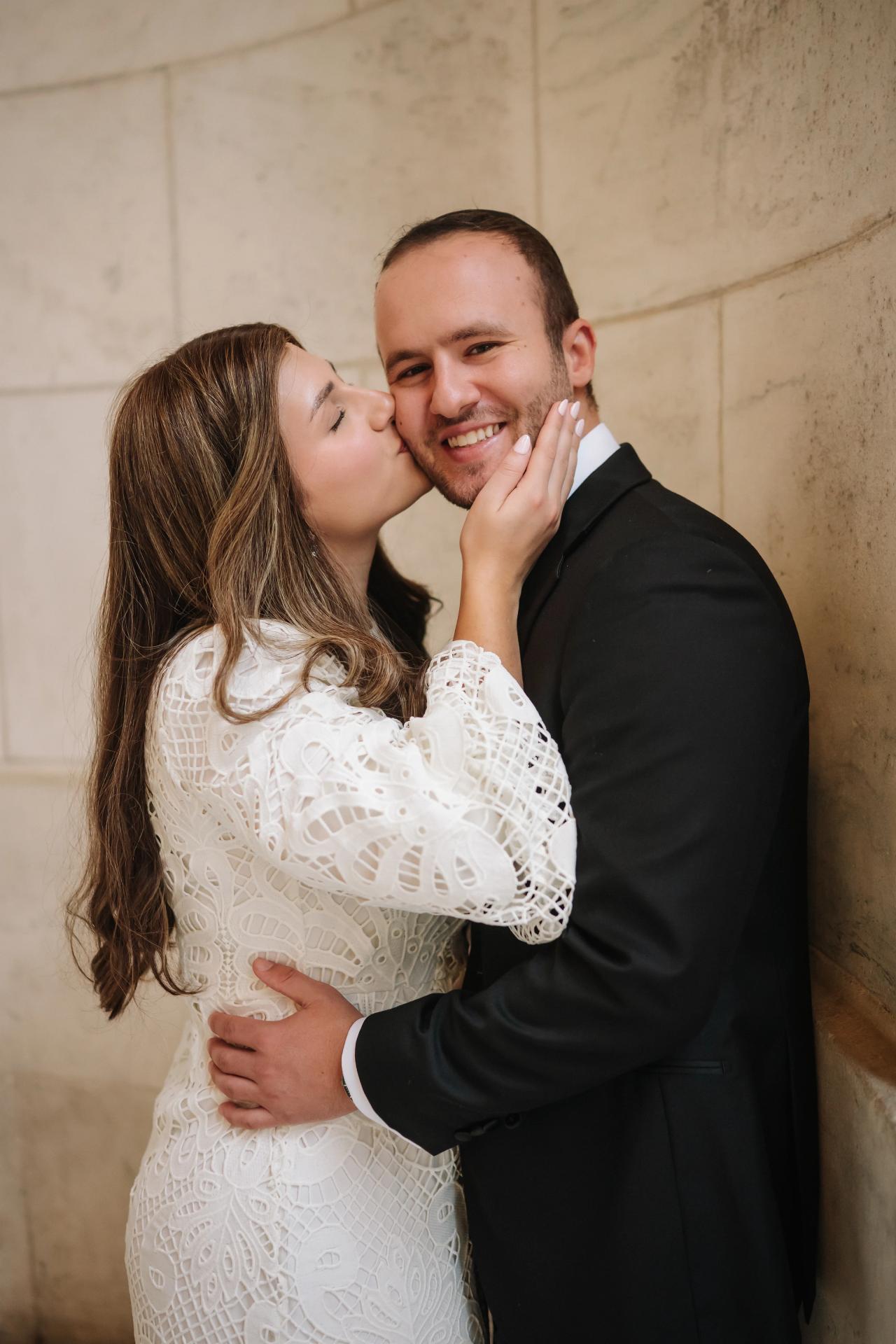 Bride and groom in a close embrace, her cheek against his, both smiling in front of a cream stone wall backdrop
