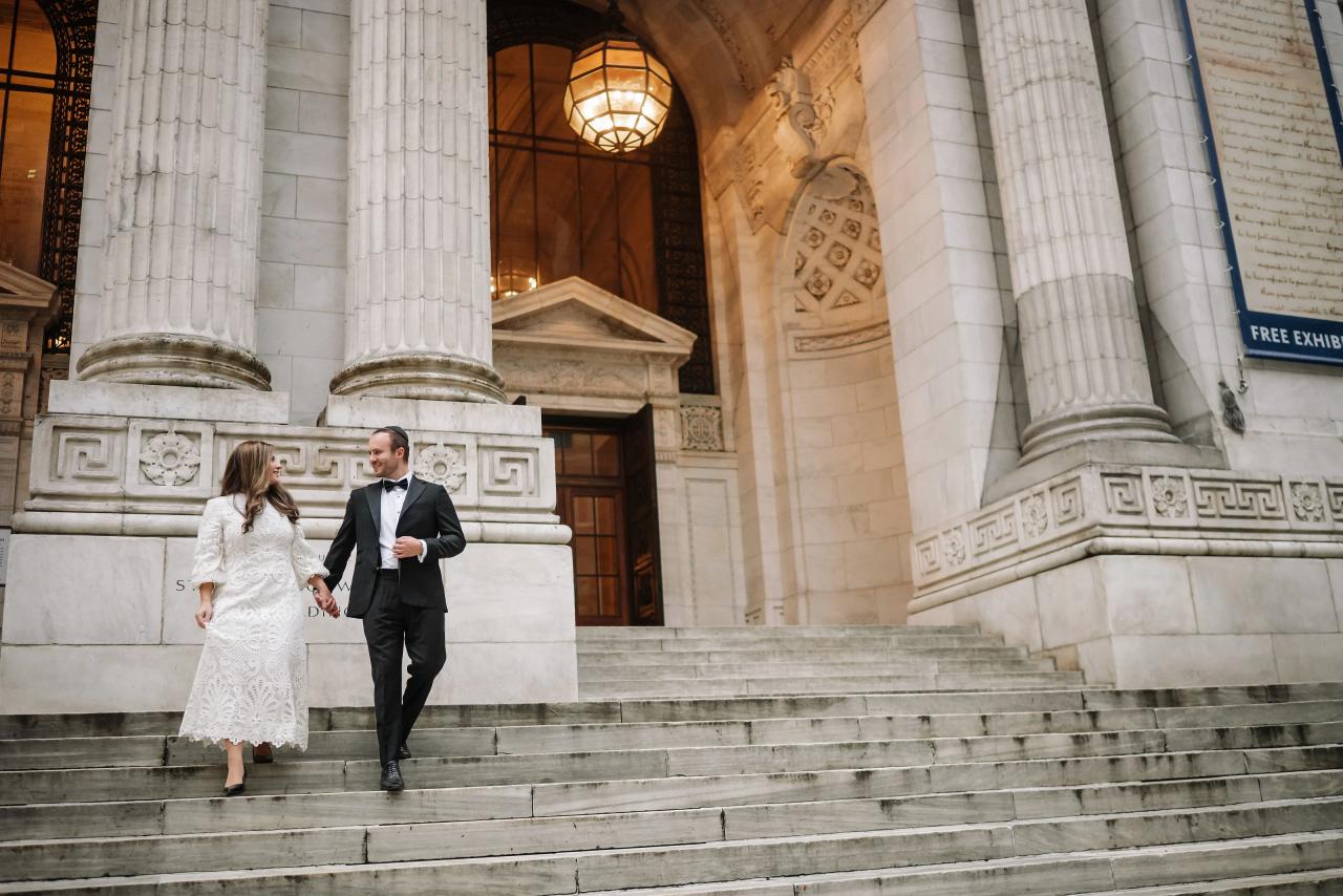 Bride in a lace dress and groom in a dark suit walking hand in hand down large stone stairs in front of classical columns and arched doors, warm interior light behind them.